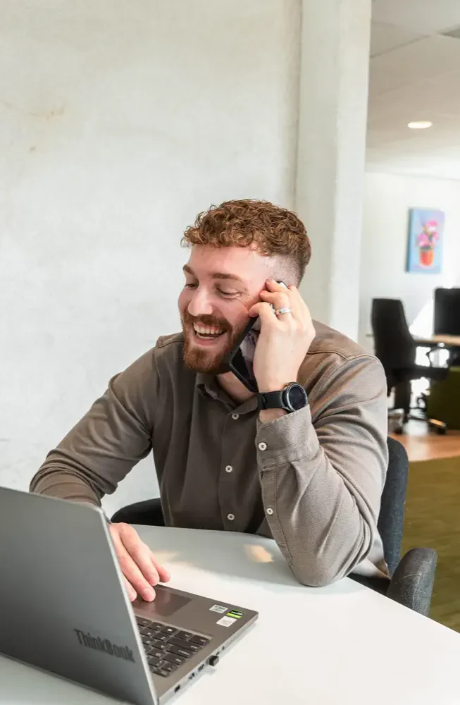 Lachende adviseur van De Vaart, adviesbureau voor het mkb, belt vanaf een werkplek achter de laptop