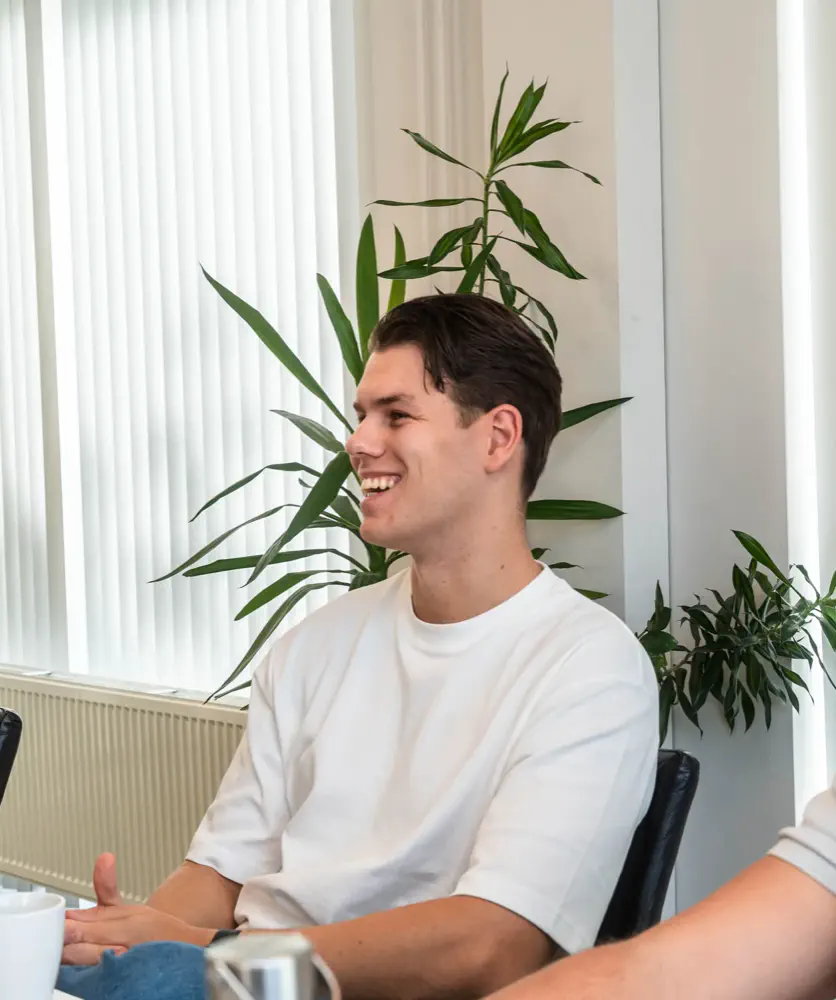 Lachende collega van De Vaart in een wit T-shirt aan de vergadertafel, met een plant op de achtergrond